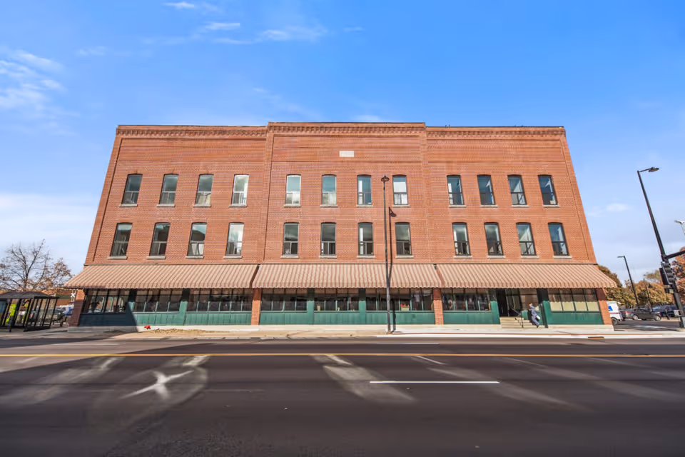 Front exterior view of a three-story red brick building with multiple windows and a striped awning covering the ground floor windows. The building is situated along a street with a sidewalk and a few pedestrians visible near the entrance.