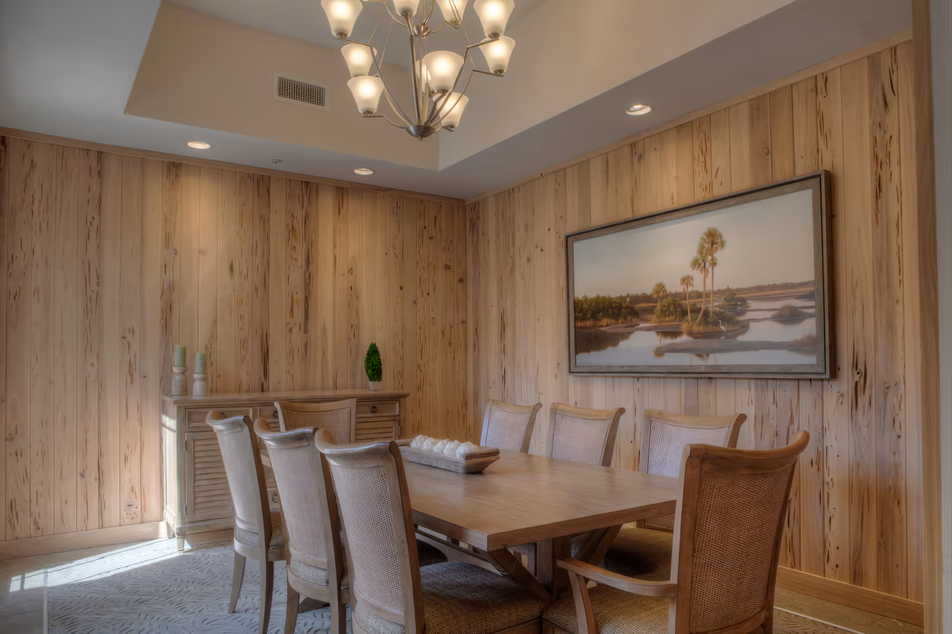 A dining room with wooden walls and a rectangular wooden dining table surrounded by eight cushioned chairs. A sideboard with decorative items is against the wall, and a large framed landscape painting hangs above it. A chandelier with multiple lights hangs from the ceiling.