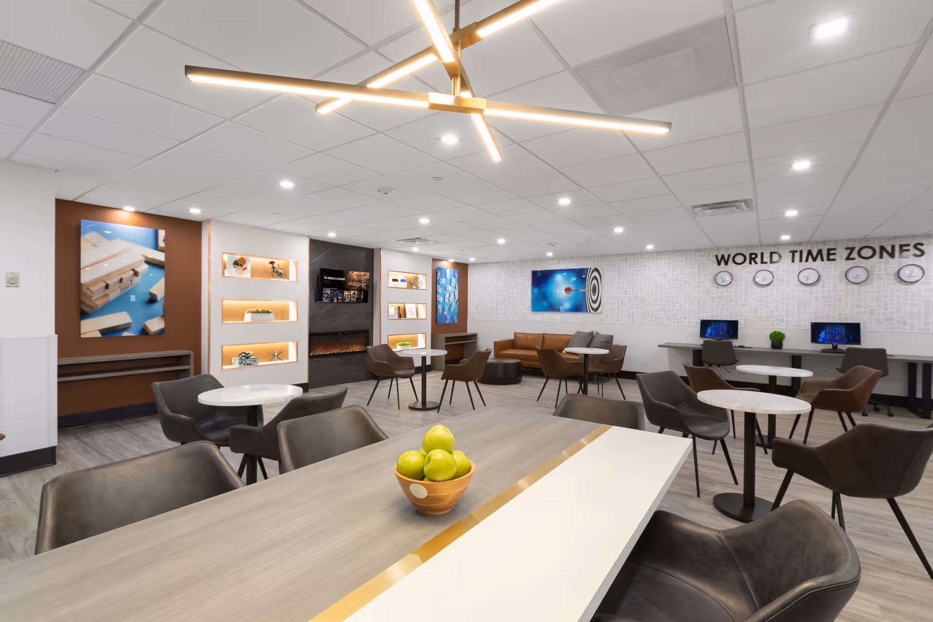 A modern common area with multiple round tables and chairs, a long table with a bowl of green apples, a brown leather couch, wall-mounted shelves with decorative items, a fireplace, and a wall displaying clocks labeled 'WORLD TIME ZONES' above computer workstations.