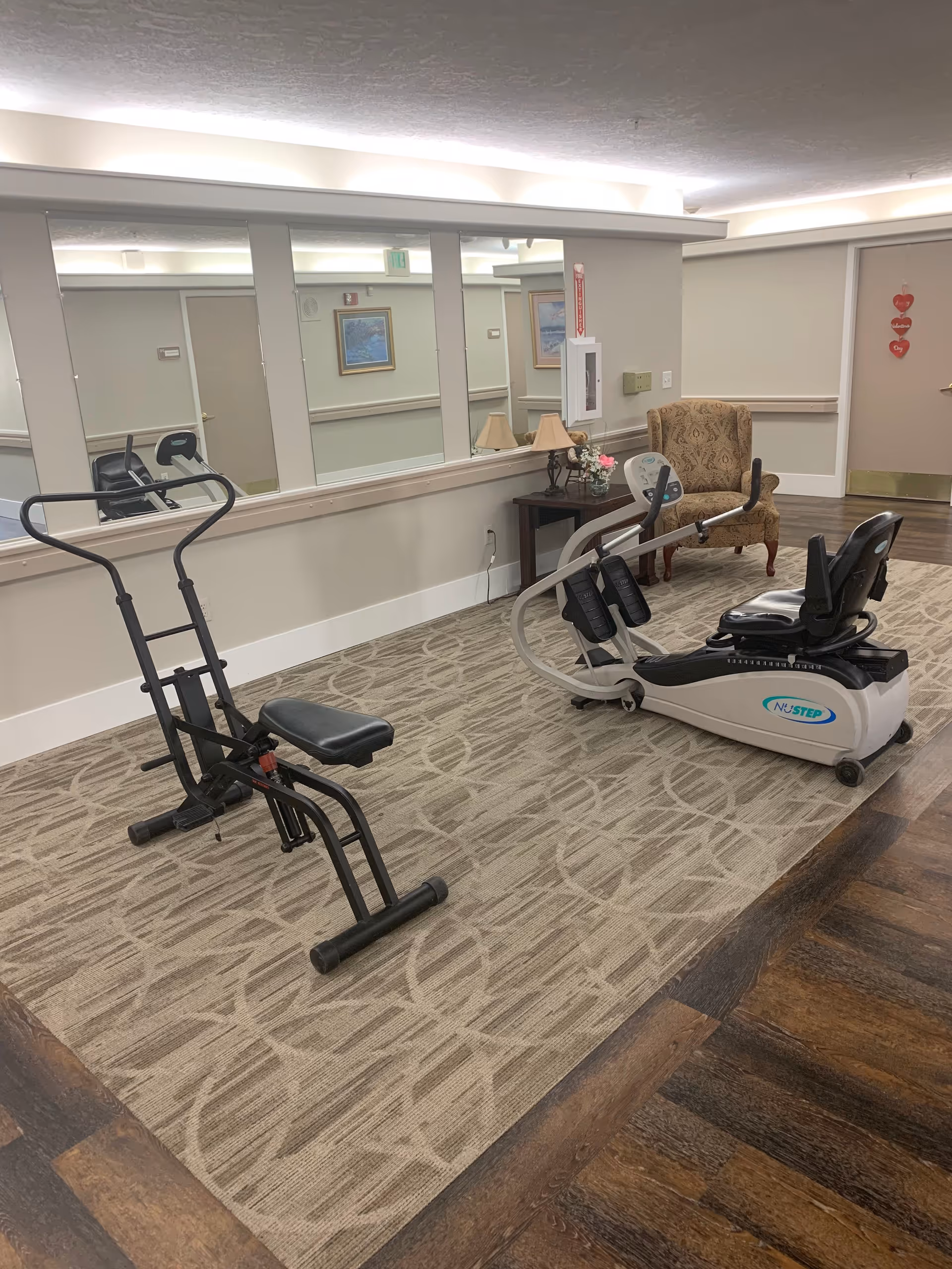 A small exercise area in Ridgeview Gardens Assisted Living featuring two pieces of exercise equipment on a patterned carpet. There is a large mirror on the wall behind the equipment, a wooden side table with a lamp and flowers, and a patterned armchair. The walls are light-colored with white trim, and there is a door decorated with three red heart-shaped ornaments.