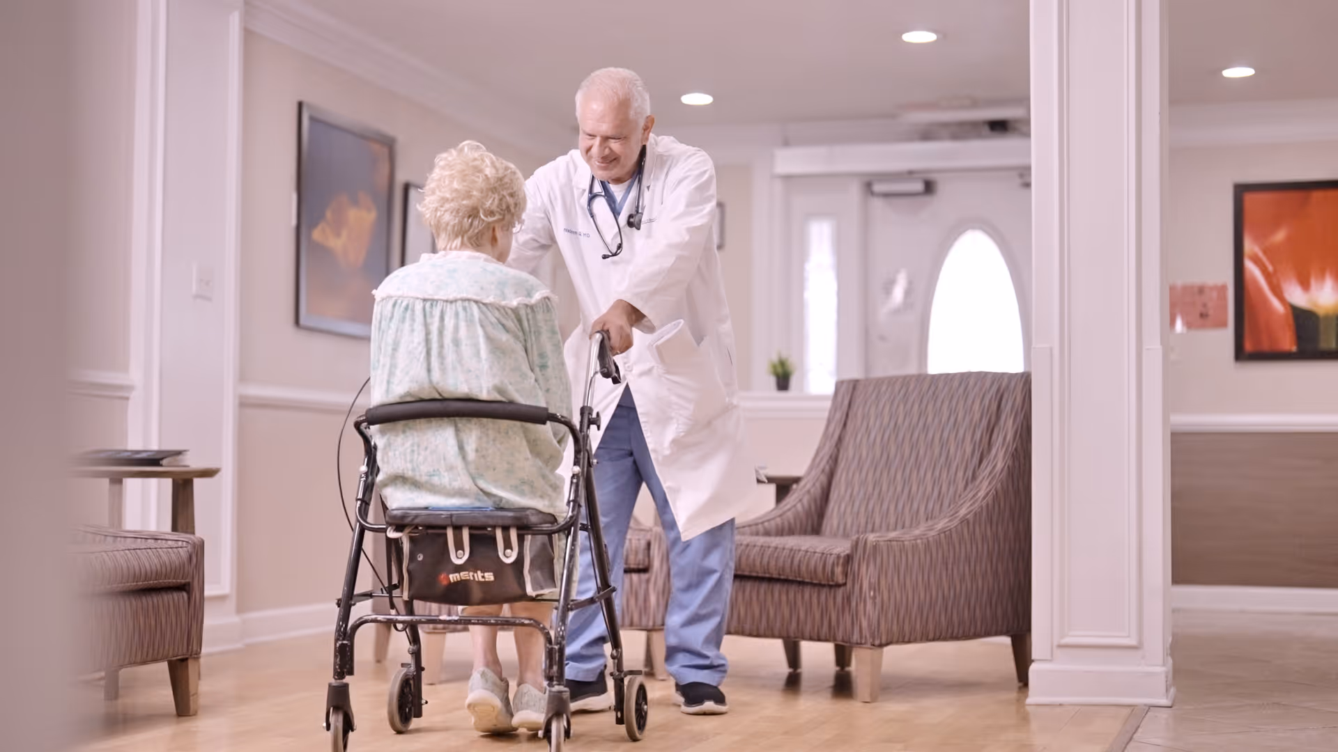 An elderly woman using a walker is assisted by a male doctor in a white coat inside a well-lit room with armchairs and framed artwork on the walls.