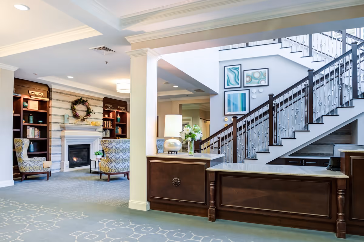 A bright and welcoming interior common area featuring a wooden reception desk with a lamp and flower vase, a staircase with decorative railing, and a cozy seating area with patterned armchairs around a fireplace. The walls are adorned with framed abstract artwork and shelves with decorative items.