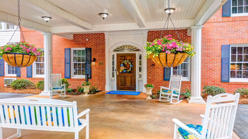 Covered entrance area of a brick building with white columns and hanging flower baskets. There are white wooden rocking chairs and benches with colorful cushions arranged on the porch. The front door is wooden with a decorative wreath and a blue welcome mat in front of it.
