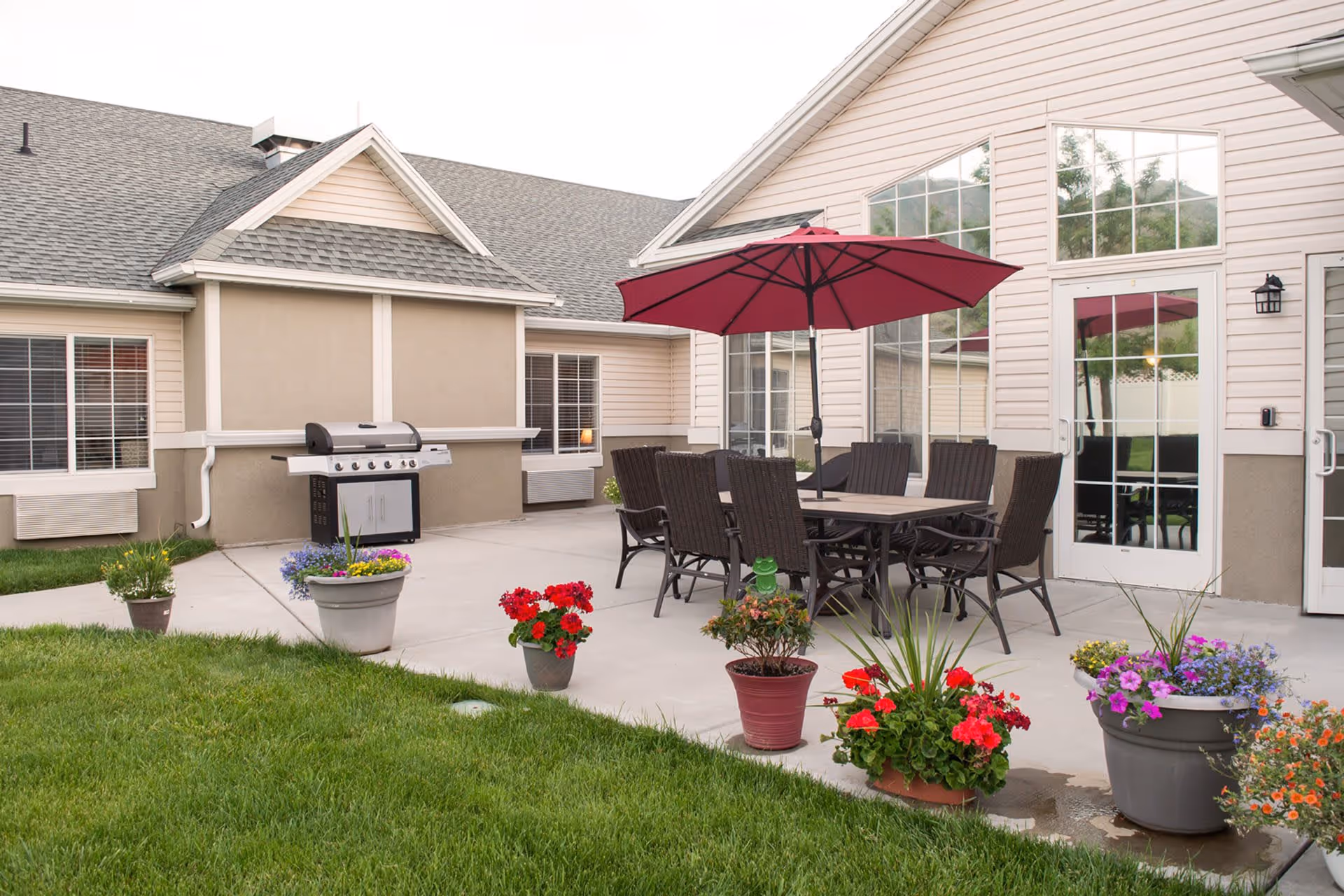 A patio with a dining table, chairs and a red umbrella surrounded by potted flowers and a gas grill outside a residential building.