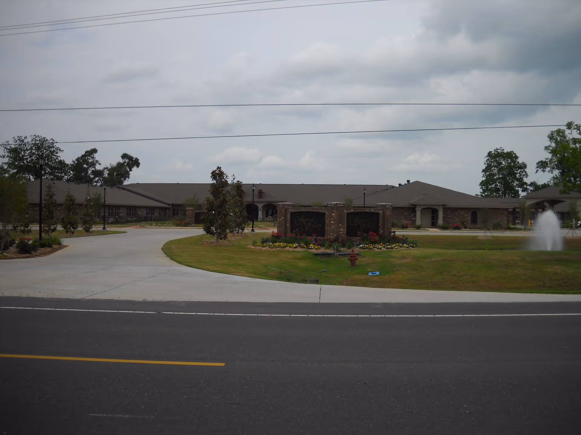 Front view of a single-story brick senior living facility with a circular driveway, landscaped lawn, and a fountain.