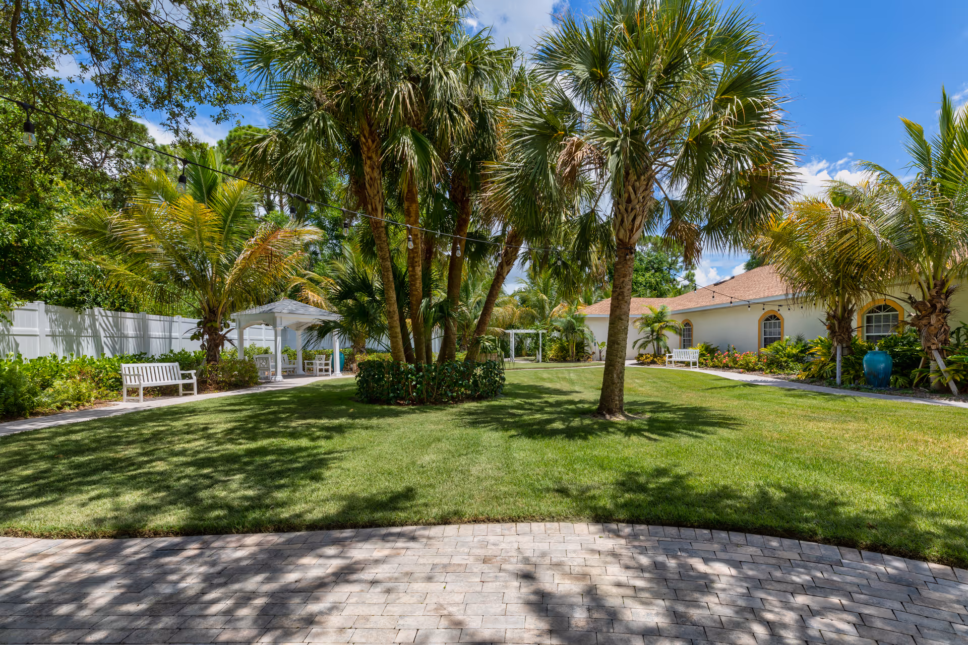 Sunny landscaped courtyard with palm trees, benches, a white gazebo, and a building in the background.
