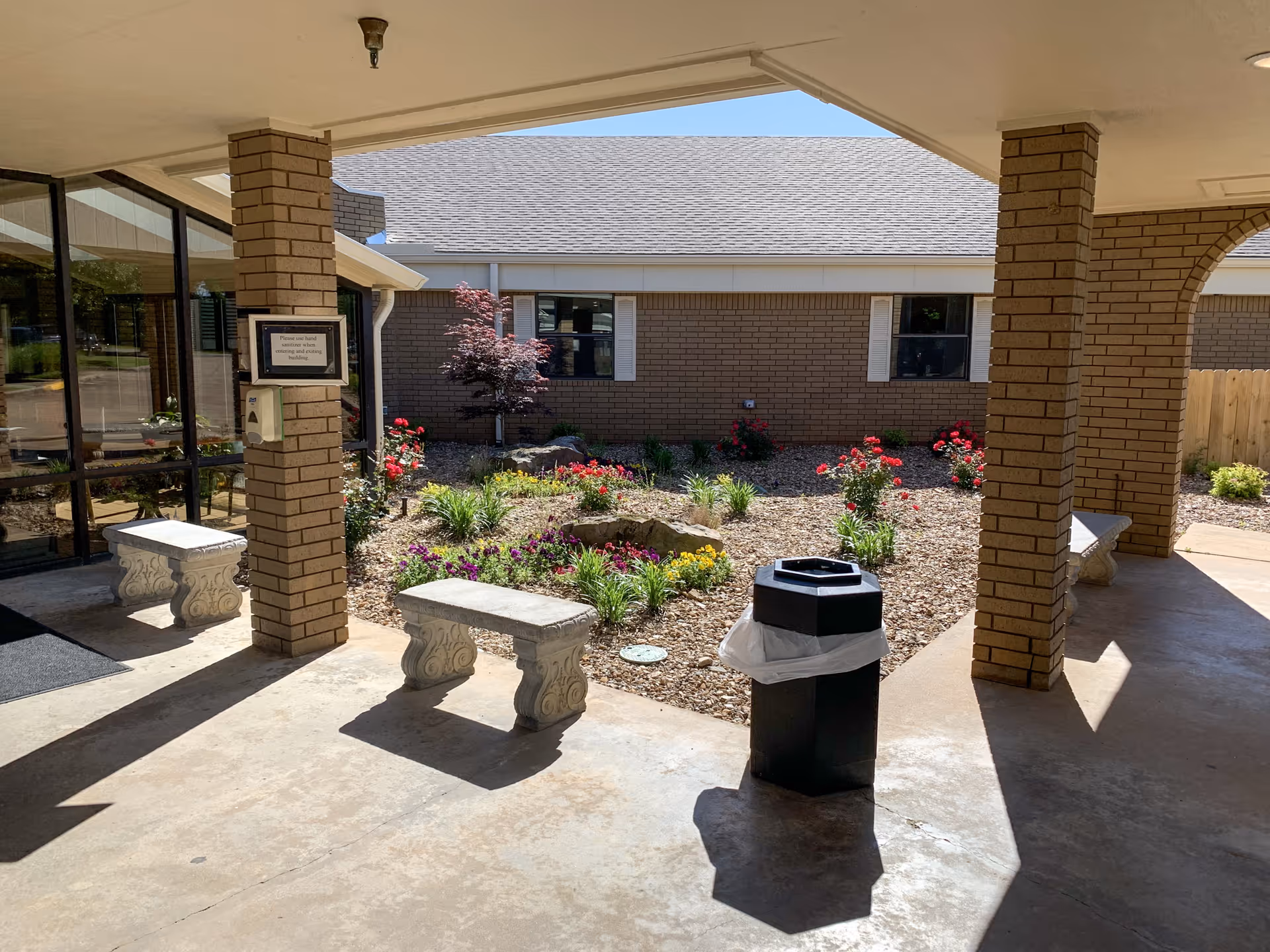 Covered outdoor seating area with stone benches and a trash can, overlooking a landscaped garden with flowers and shrubs in front of a brick building under a clear blue sky.