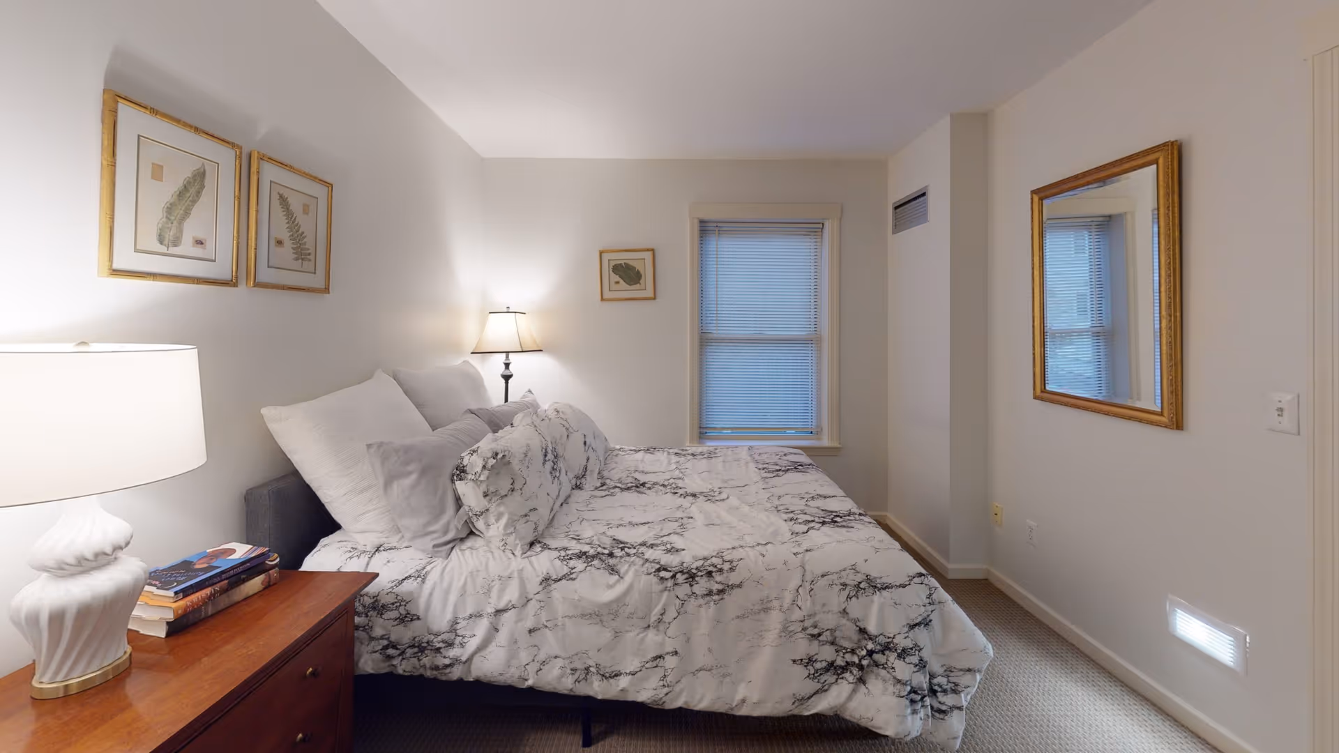 Bright, neutral bedroom with a bed covered in marble-patterned bedding, a nightstand with a lamp and books, framed artwork on the walls and a mirror.