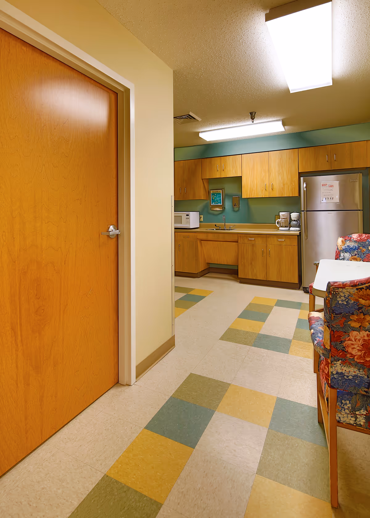 Small kitchenette and seating area with wood cabinets, a stainless refrigerator, microwave and sink, patterned tile floor and floral-upholstered chairs.