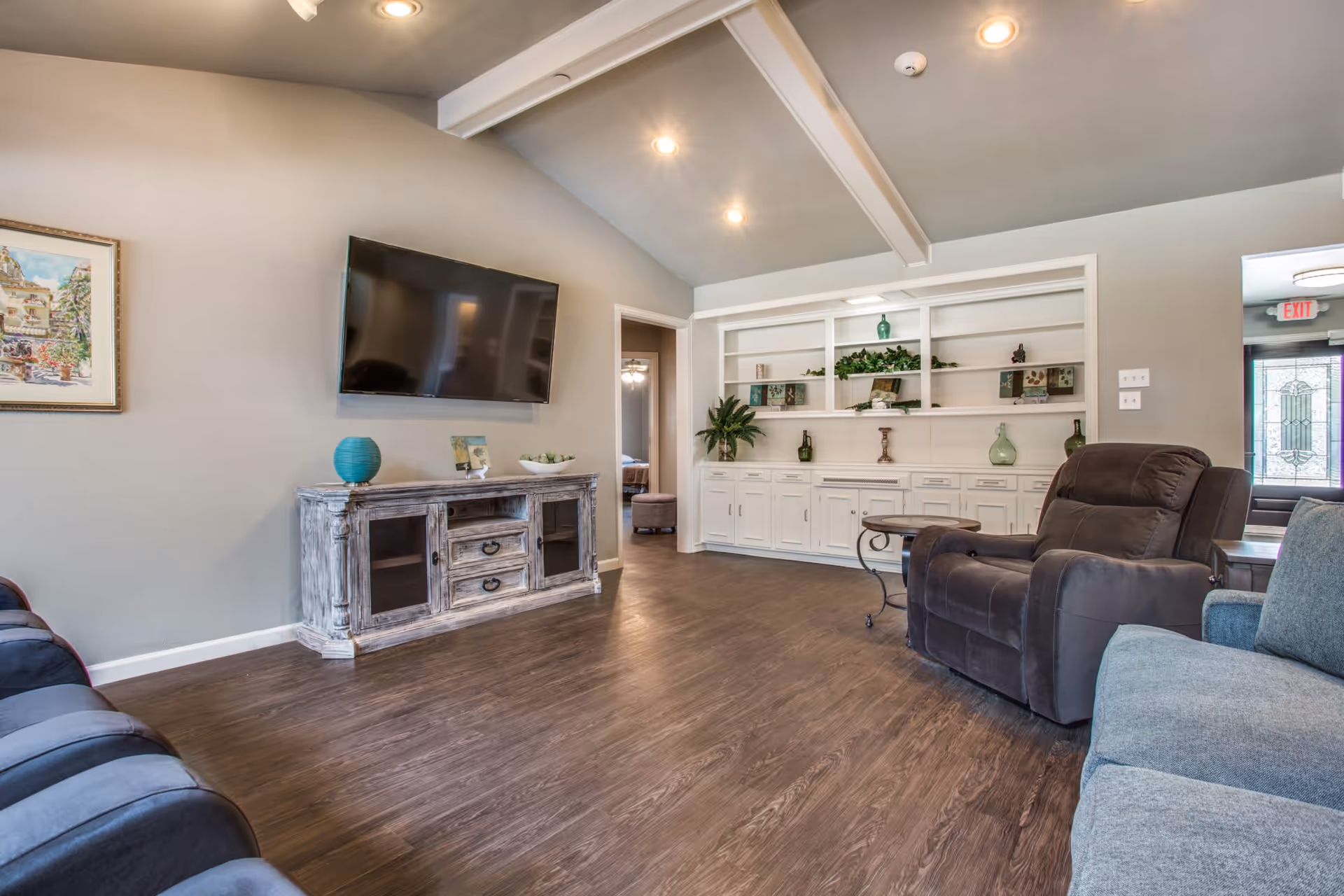 A cozy living room with a mounted flat-screen TV above a rustic wooden cabinet, a dark brown recliner, a blue sofa, a small round side table, and built-in white shelves decorated with plants and vases. The room has wooden flooring, recessed ceiling lights, and a doorway leading to another room with a bed visible.
