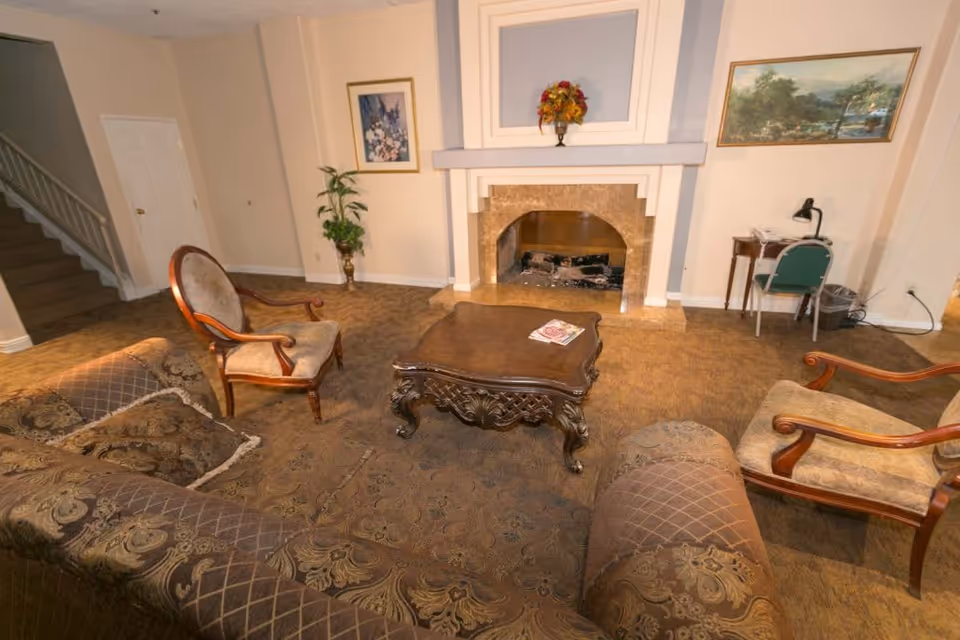 A cozy living room with a patterned brown carpet, a brown patterned sofa, two wooden armchairs with beige cushions, and a carved wooden coffee table with a magazine on it. There is a fireplace with a floral arrangement on the mantel, a potted plant in the corner, a small desk with a green chair and a lamp, and framed artwork on the walls.