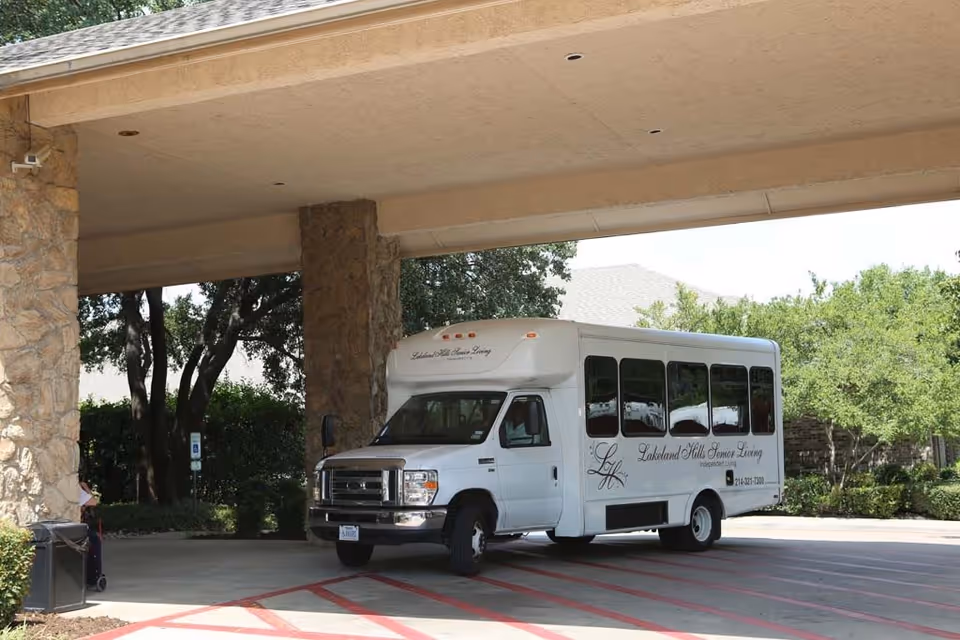 A white shuttle bus parked under a covered driveway area with stone pillars. The bus has the text 'Lakeland Hills Senior Living' and a phone number on its side. Trees and greenery are visible in the background.
