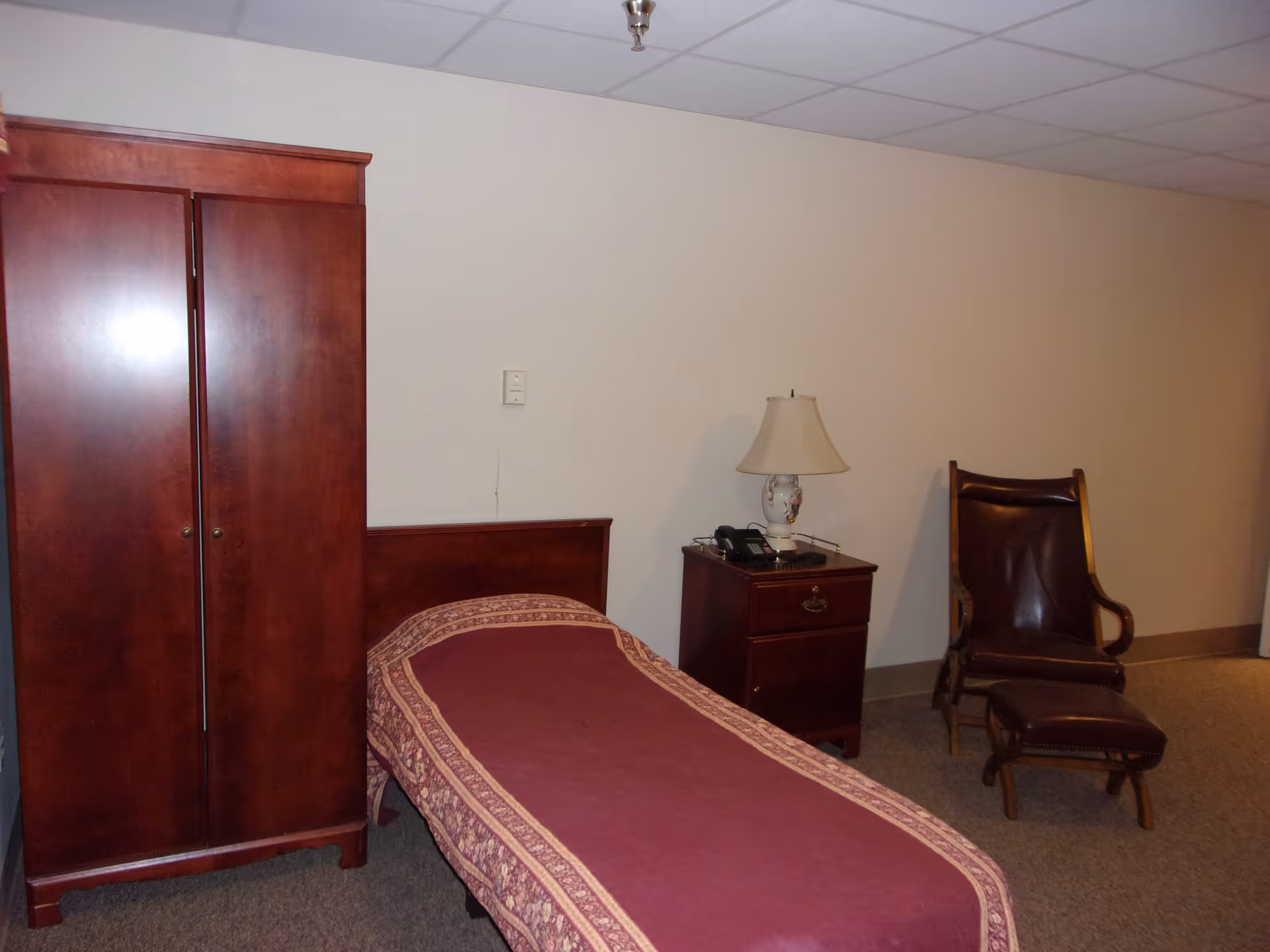 A simple bedroom with a single bed covered in a maroon and patterned bedspread, a wooden wardrobe, a wooden nightstand with a decorative lamp and a telephone, and a wooden chair with a matching footrest against a plain beige wall.
