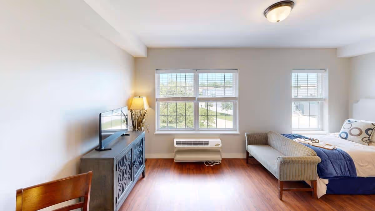 A bright and clean senior living bedroom with a bed, a small sofa, a wooden chair, a TV on a decorative cabinet, a lamp, and large windows letting in natural light. The room has light-colored walls and wooden flooring.