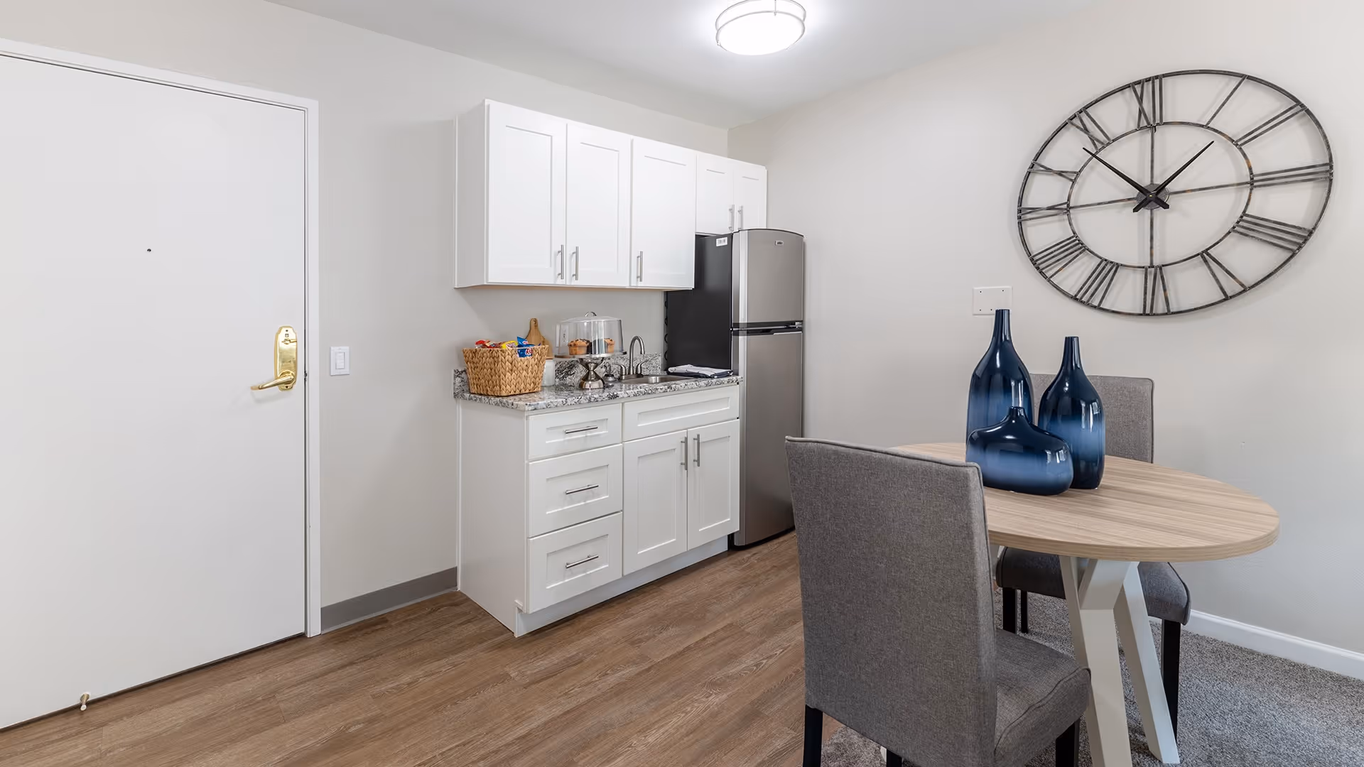 Small kitchenette area with white cabinets, a granite countertop, a stainless steel mini refrigerator, and a basket with snacks. Next to the kitchenette is a small round wooden dining table with two gray upholstered chairs and three decorative blue vases on top. A large wall clock with Roman numerals hangs on the wall above the table. The floor is wood laminate and there is a white door to the left.