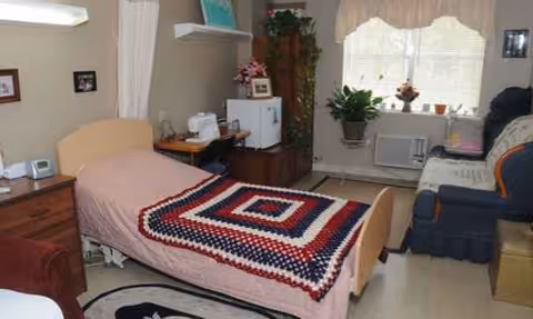 Bedroom in a senior living facility with a bed, crochet blanket, chair, and windowsill plants.