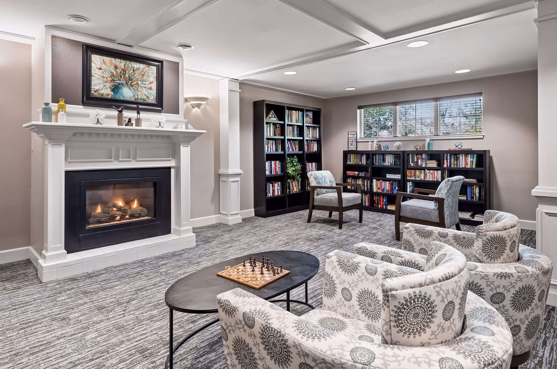 A cozy living room area with a lit fireplace on the left, a framed floral painting above it, and two black bookshelves filled with books against the back wall. There are four patterned armchairs arranged around a black oval coffee table with a chessboard on it. The room has a large window with blinds letting in natural light and neutral-colored walls and carpet.