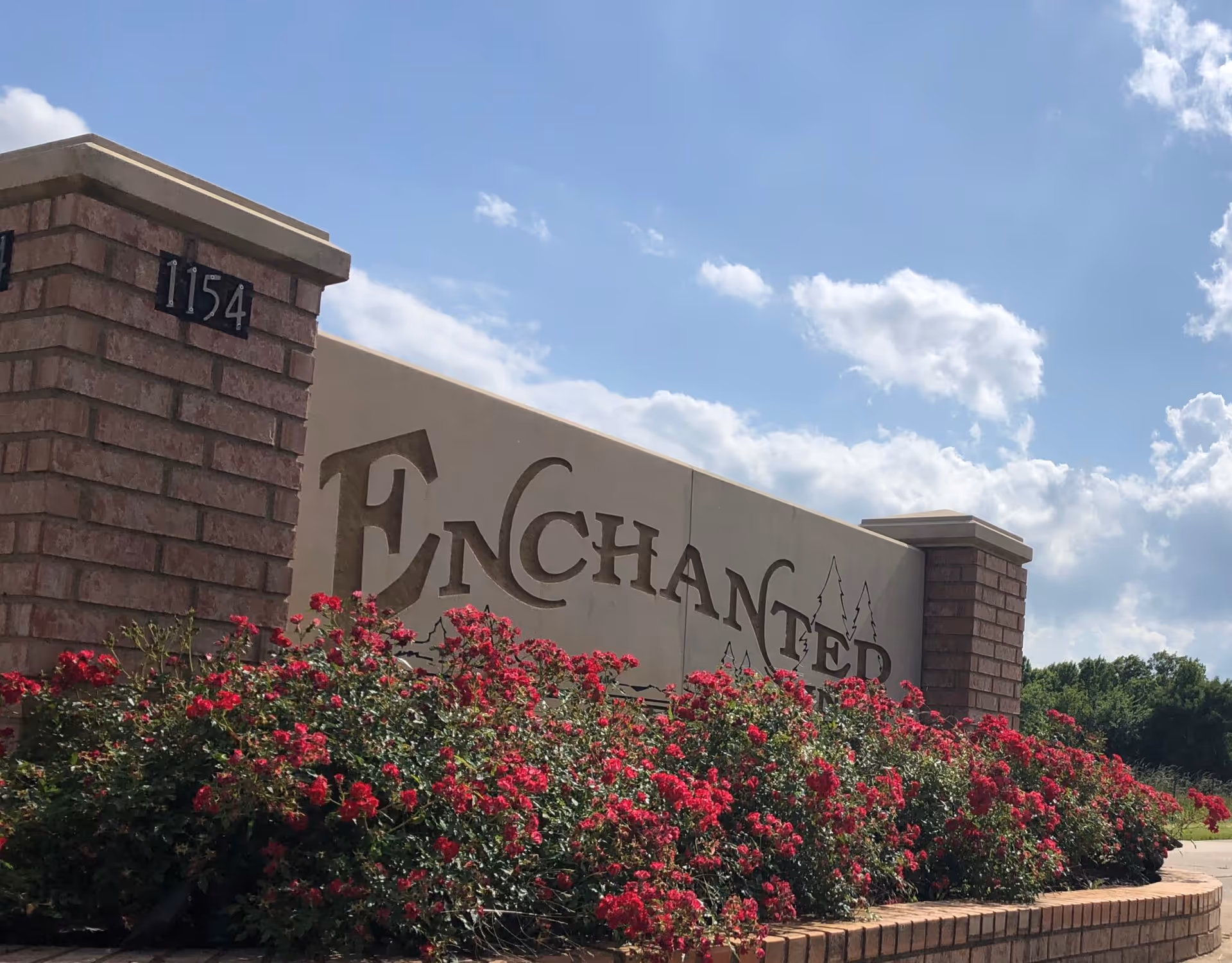 A brick and stone entrance sign for Enchanted Pines facility with the address number 1154 displayed on a brick pillar. The sign is surrounded by vibrant red flowers and greenery under a partly cloudy blue sky.
