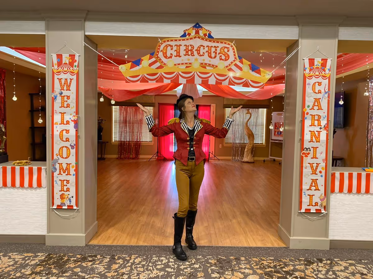 A woman dressed as a ringmaster stands with arms raised under a 'CIRCUS' sign in a decorated indoor room with 'WELCOME' and 'CARNIVAL' banners.
