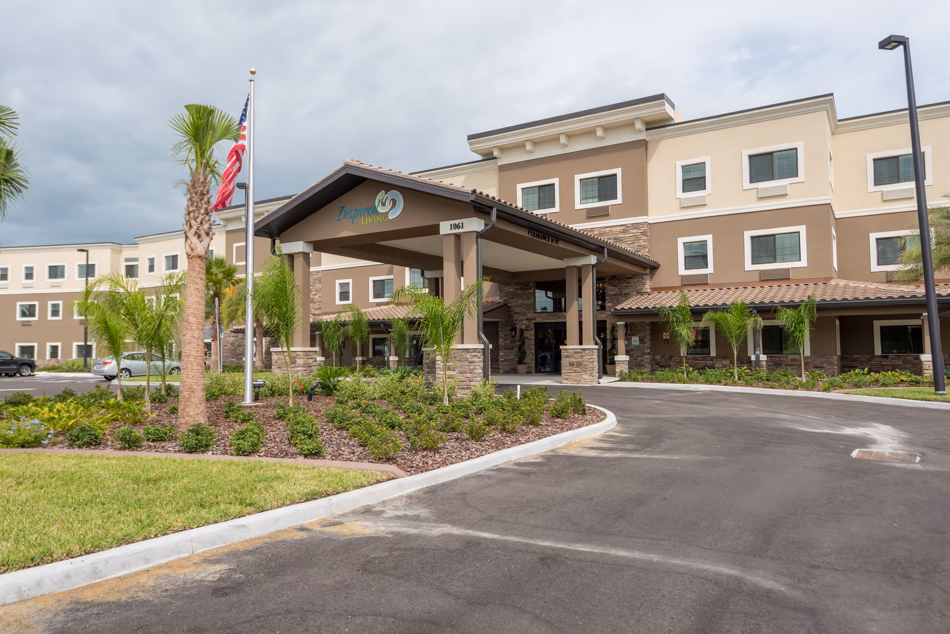 Exterior view of Inspired Living Ocoee, a multi-story senior living facility with a covered entrance, landscaped garden with palm trees, and an American flag on a flagpole in front.