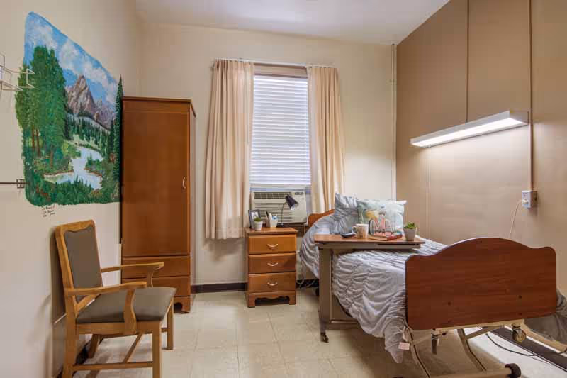 A simple nursing facility bedroom with a single hospital bed, a wooden bedside table with drawers, a wooden wardrobe, and a cushioned wooden chair. The bed has a gray comforter and pillows, and there is a small table on wheels positioned over the bed with a mug and a small plant. A window with beige curtains and a window air conditioning unit is centered on the back wall. A landscape mural depicting trees, mountains, and a river is painted on the left wall.