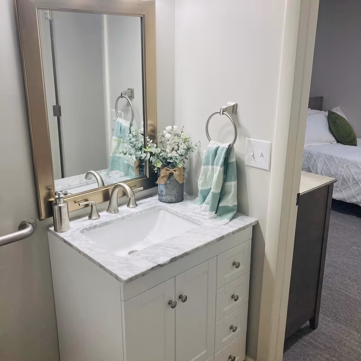 A bathroom vanity with a white marble countertop, a rectangular sink, and a silver faucet. Above the sink is a large mirror with a gold frame. On the countertop, there is a small metal vase with green and white artificial flowers and a soap dispenser. A green and white striped hand towel hangs on a silver towel ring mounted on the wall. To the right, there is a doorway leading to a bedroom with a bed that has white bedding and a green pillow.