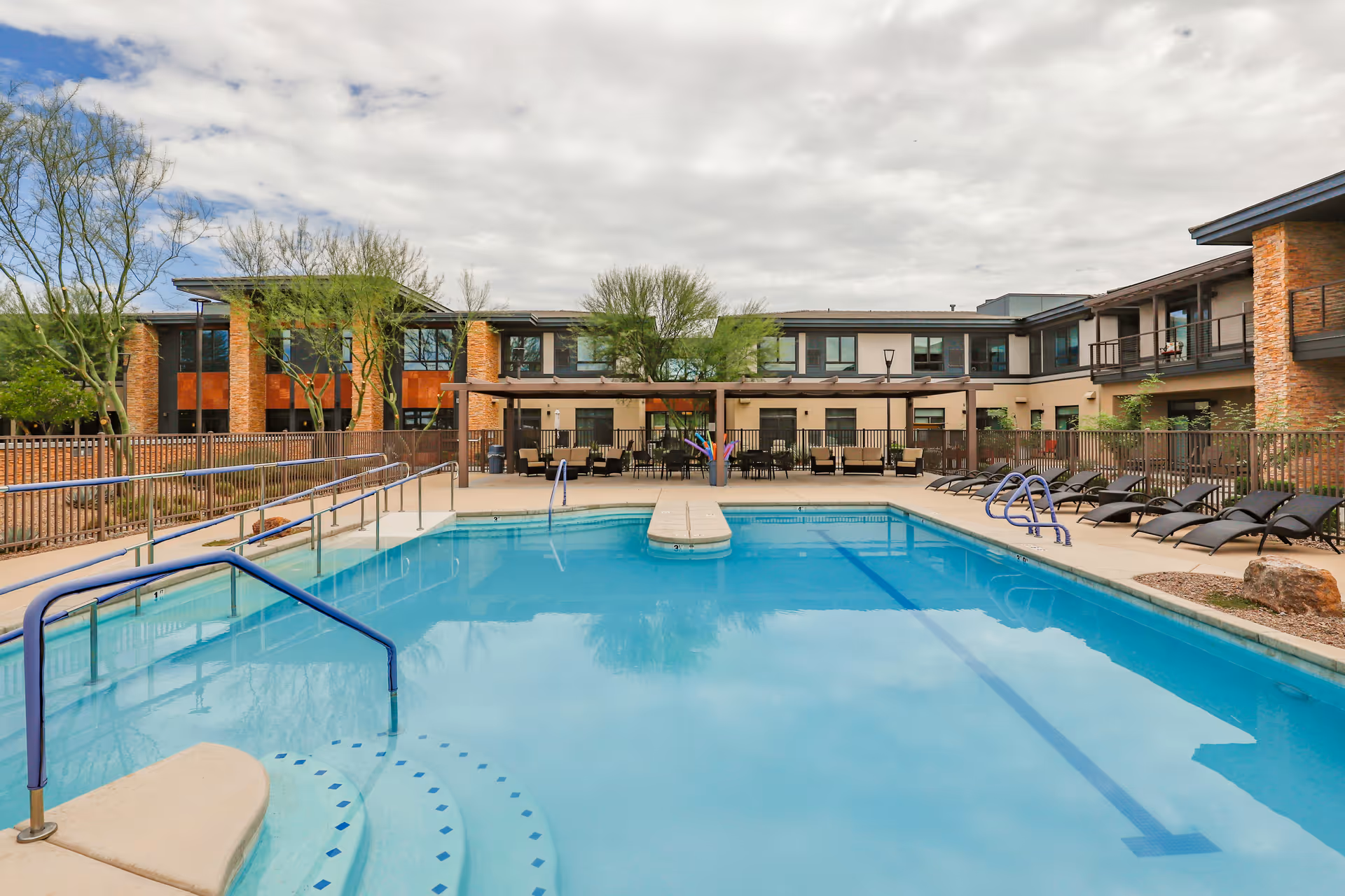 Outdoor swimming pool with handrails and steps leading into the water, surrounded by lounge chairs and a covered seating area. The pool area is enclosed by a fence, and a two-story building with large windows and brick accents is visible in the background under a cloudy sky.