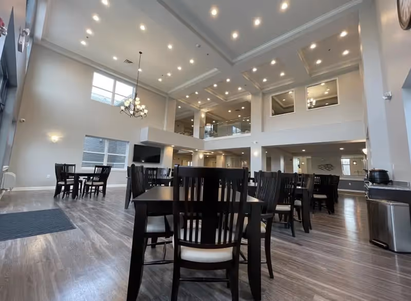 Spacious dining area with multiple dark wooden tables and chairs arranged on a wood-patterned floor. The room features high ceilings with recessed lighting, large windows allowing natural light, a chandelier, and a mounted TV on the far wall. There is a second-floor balcony overlooking the dining area.