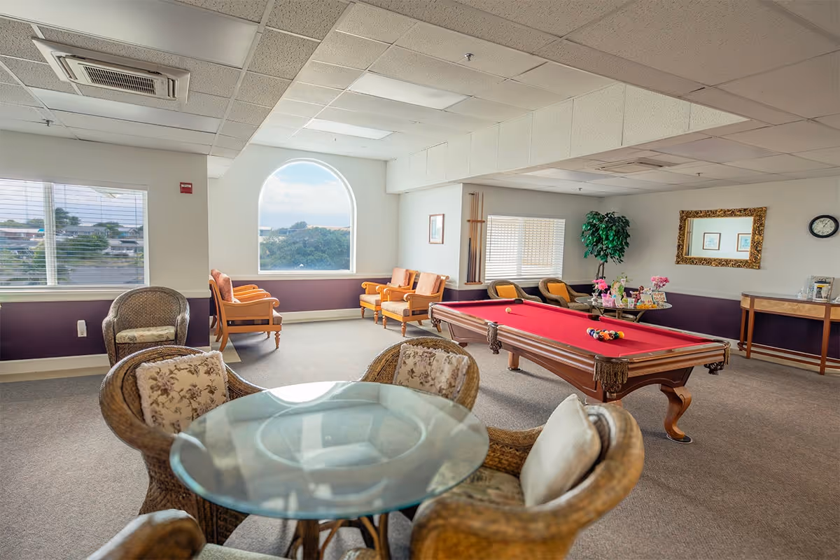 A bright and spacious common room with a red pool table in the center, surrounded by several cushioned chairs and wicker seating. The room features large windows, including an arched window, allowing natural light to fill the space. There is a glass-top round table with wicker chairs in the foreground, a wall clock, a large mirror, and a small table with decorative items against the wall.