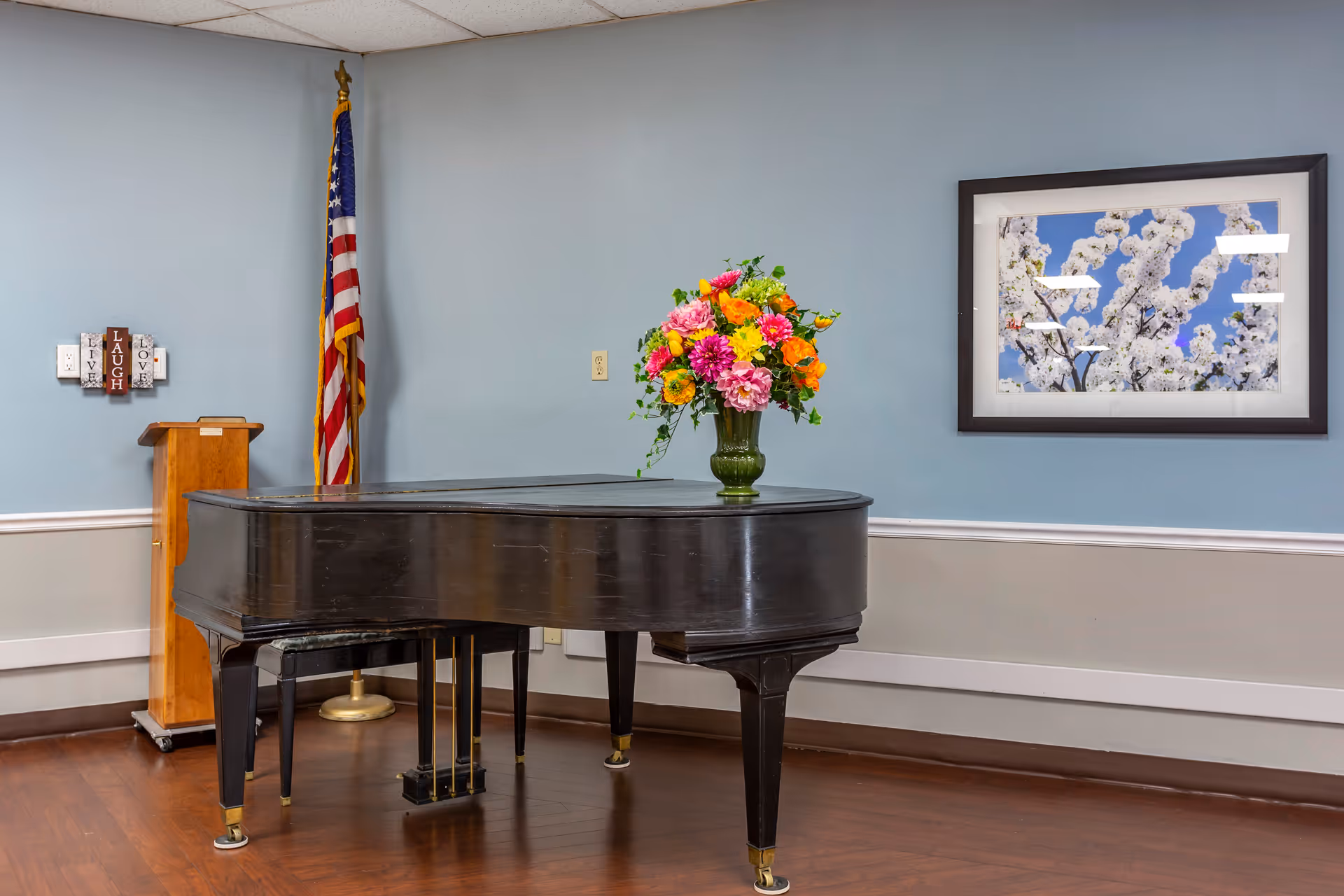 A black grand piano with a colorful flower arrangement on top is placed in a room with light blue walls and wooden flooring. An American flag stands in the corner next to a wooden podium, and a framed picture of white blossoms against a blue sky hangs on the wall.
