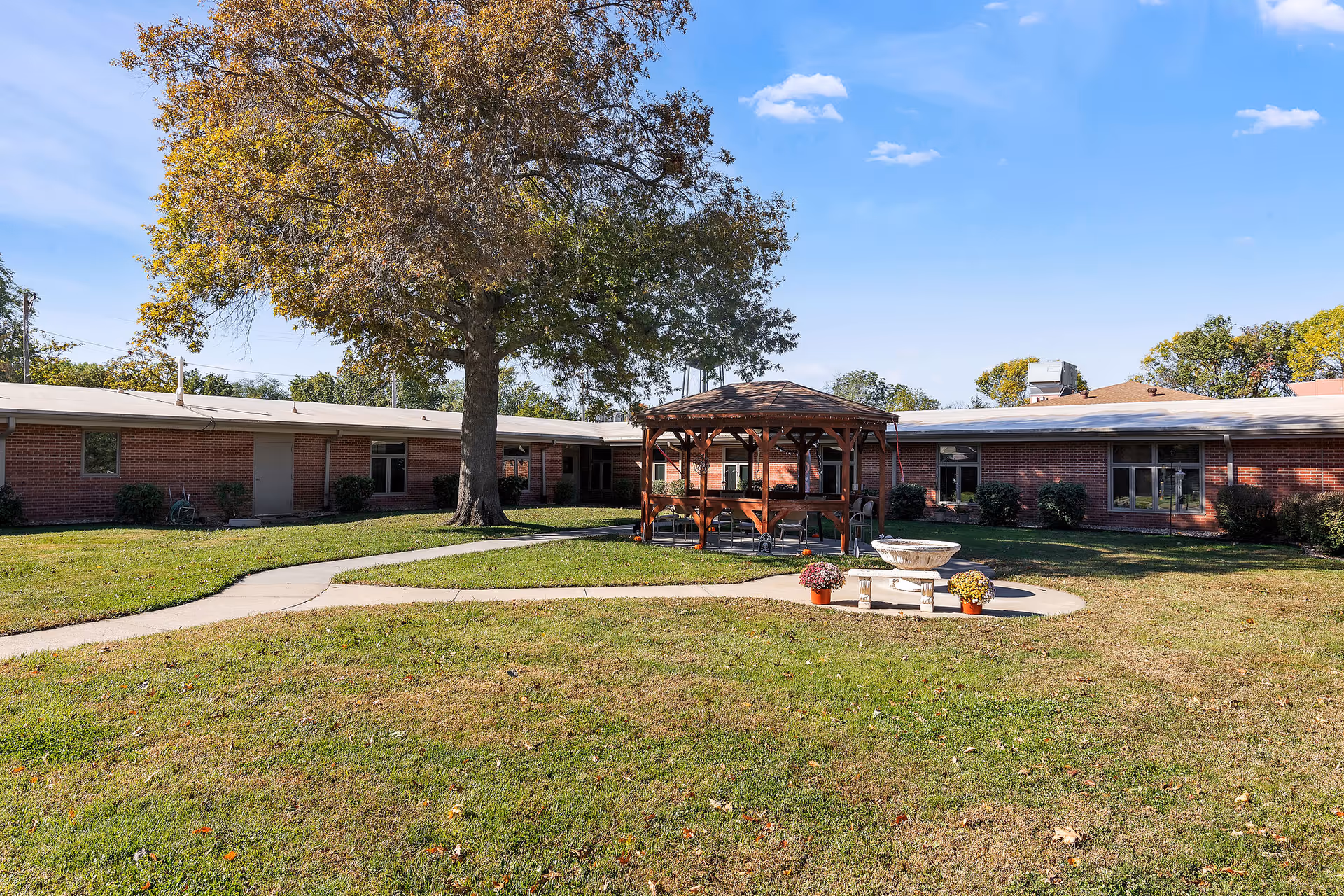 Outdoor courtyard area of Parkview Heights Care Center featuring a large tree, a wooden gazebo with seating underneath, a concrete pathway, and a decorative stone fountain surrounded by potted flowers. The courtyard is enclosed by a single-story brick building under a clear blue sky.