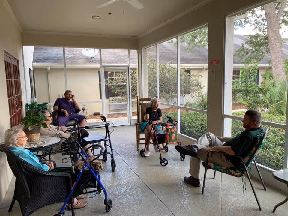 A group of elderly people and a caregiver sitting and relaxing in a screened-in porch area with large windows overlooking a garden. Some individuals are seated in chairs, and some have walkers nearby. The setting appears calm and social.
