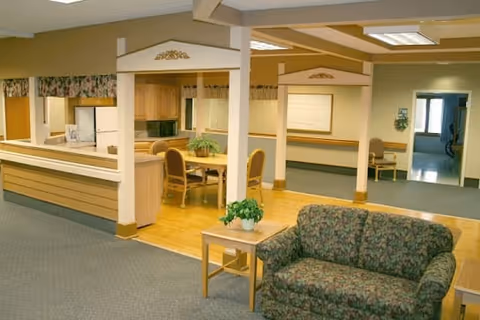 Interior view of a senior living facility showing a common area with a floral patterned couch and a small wooden table with a potted plant. In the background, there is a dining area with a wooden table and chairs, a kitchen area with appliances, and a hallway leading to other rooms.