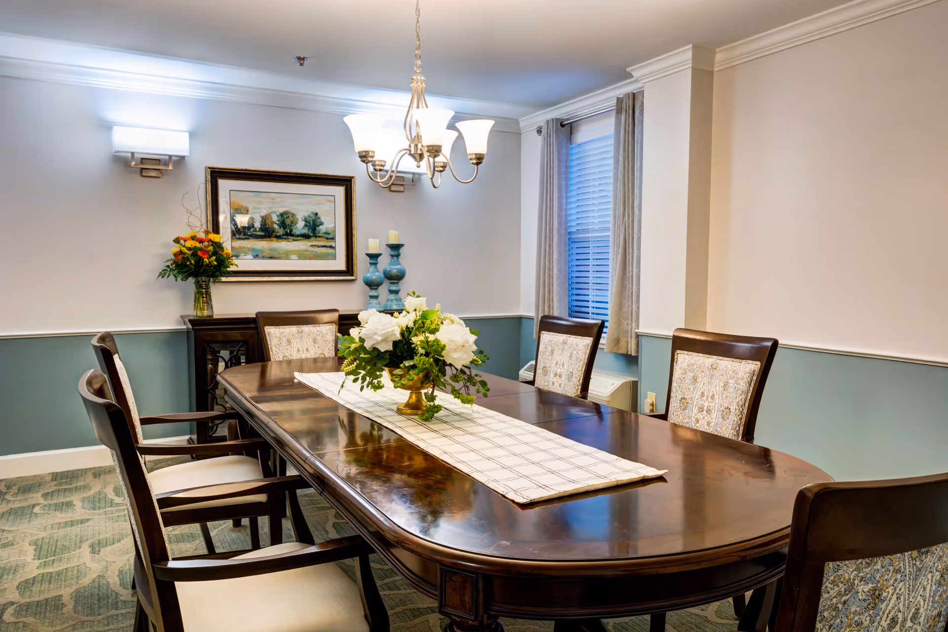 A formal dining room with a long polished wooden table surrounded by six upholstered chairs. A white and beige table runner with a floral centerpiece is placed on the table. The room has light blue and white walls, a window with beige curtains, a chandelier hanging from the ceiling, a framed landscape painting on the wall, and a sideboard with decorative items including a vase of flowers and two blue candle holders.