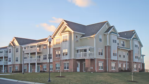 Exterior view of a multi-story senior living facility building with beige siding and brick accents, multiple balconies, and a well-maintained lawn under a partly cloudy sky.