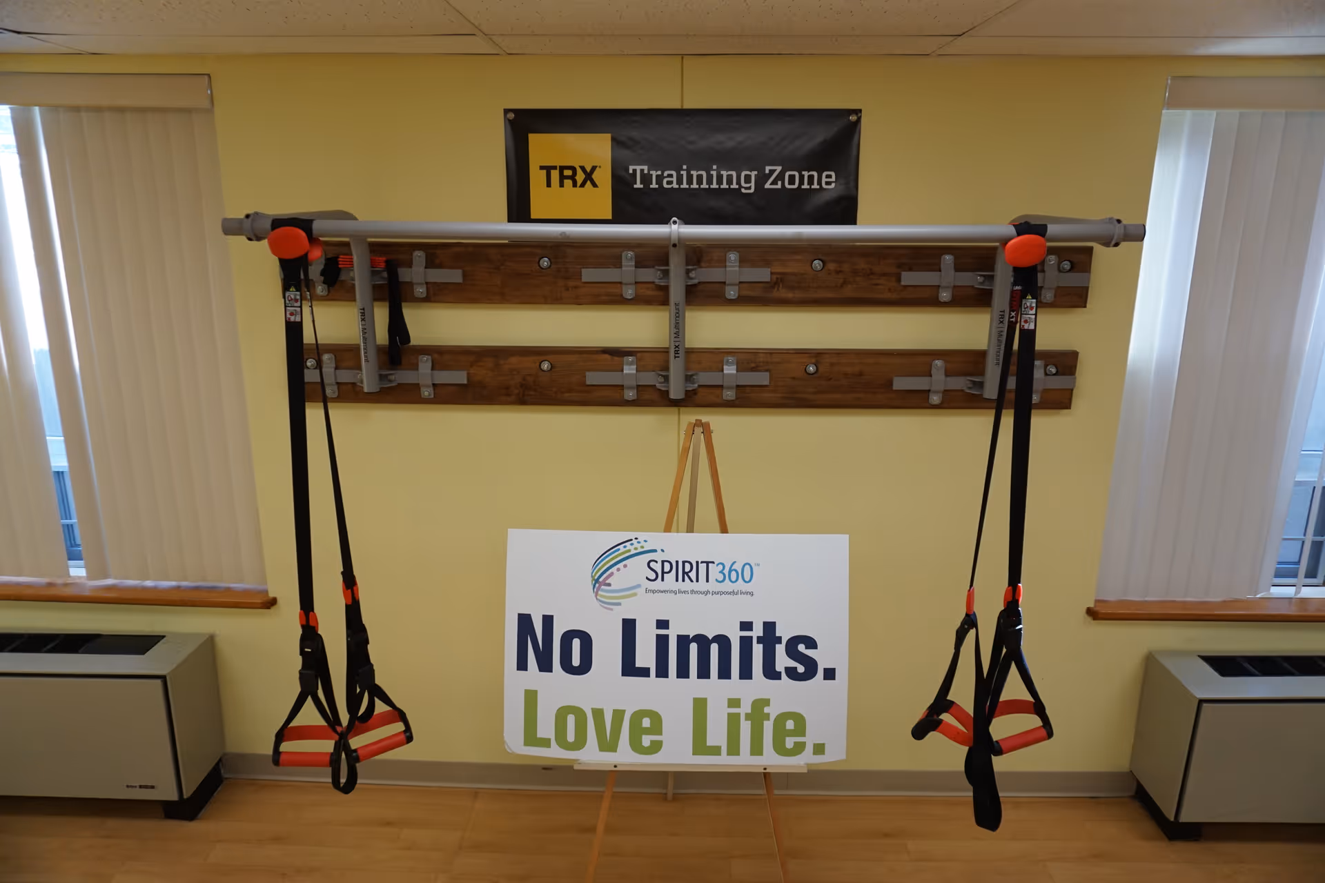 A fitness room wall with TRX suspension straps and a sign reading "No Limits. Love Life." beneath a "TRX Training Zone" banner.