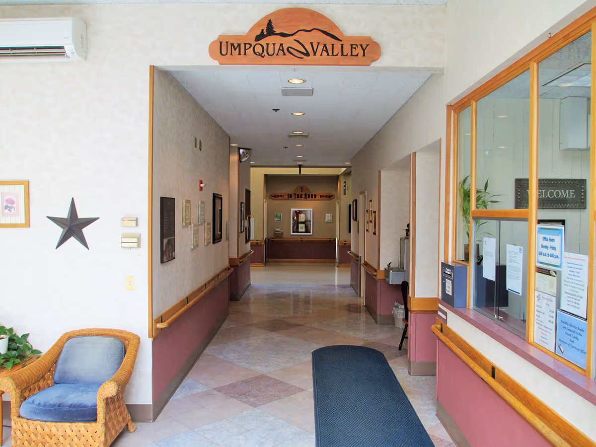 Entrance lobby and hallway of Umpqua Valley Nursing & Rehabilitation Center showing a reception window, seating area, and a long corridor.