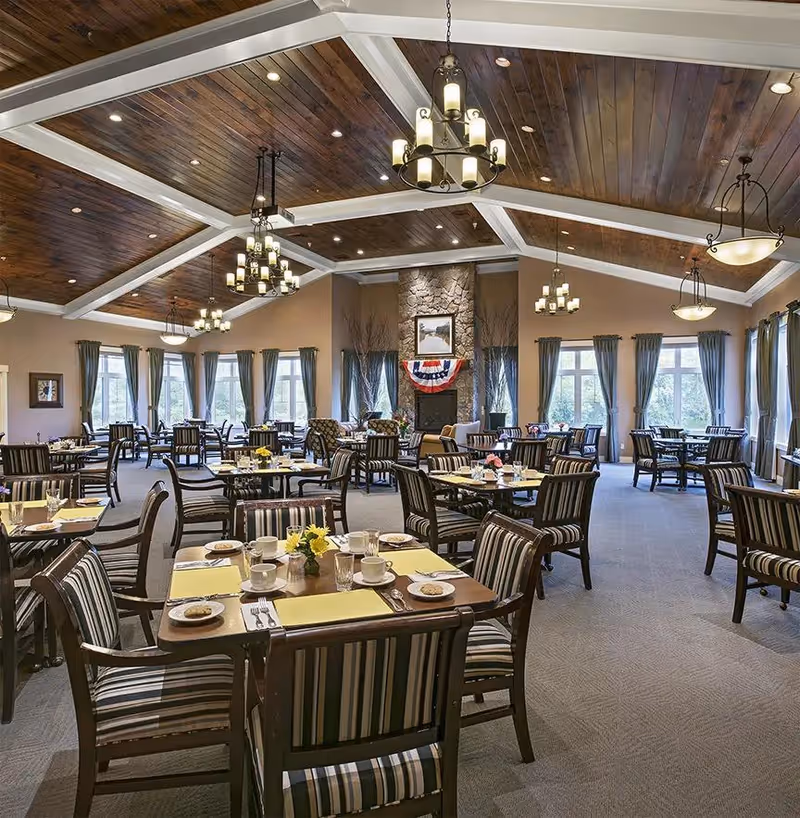 Spacious dining room with multiple tables set for meals, featuring striped cushioned chairs, yellow placemats, and plates with cookies. The room has large windows with blue curtains, a stone fireplace decorated with a patriotic banner, and a wooden vaulted ceiling with multiple chandeliers providing warm lighting.