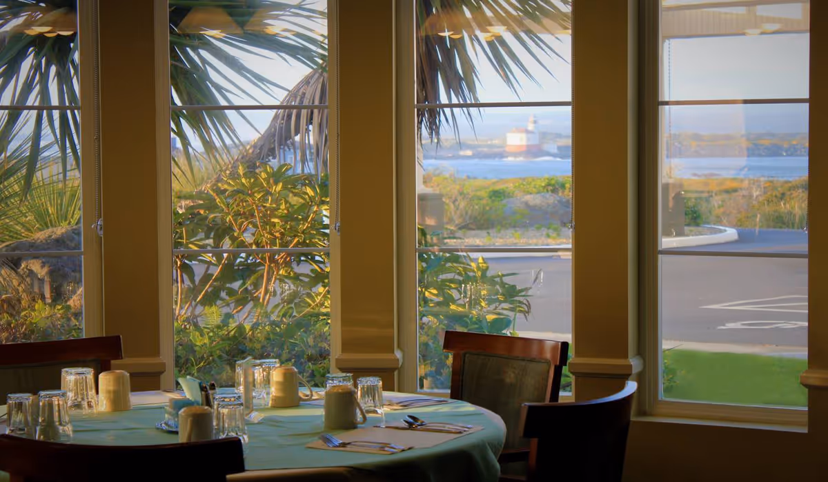 A dining table set with glasses, cups, and utensils in front of large windows showing an outdoor view with greenery, a road, and a lighthouse in the distance.