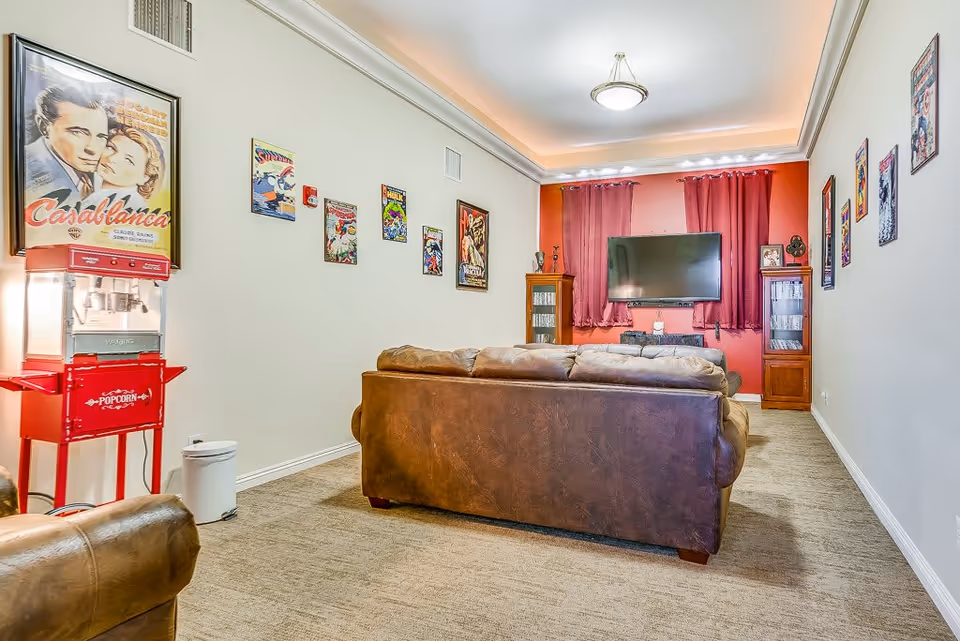 A cozy living room with a brown leather couch facing a wall-mounted flat screen TV. The room has red curtains, a popcorn machine, and vintage movie posters and comic book covers decorating the walls. There are two wooden display cabinets on either side of the TV.