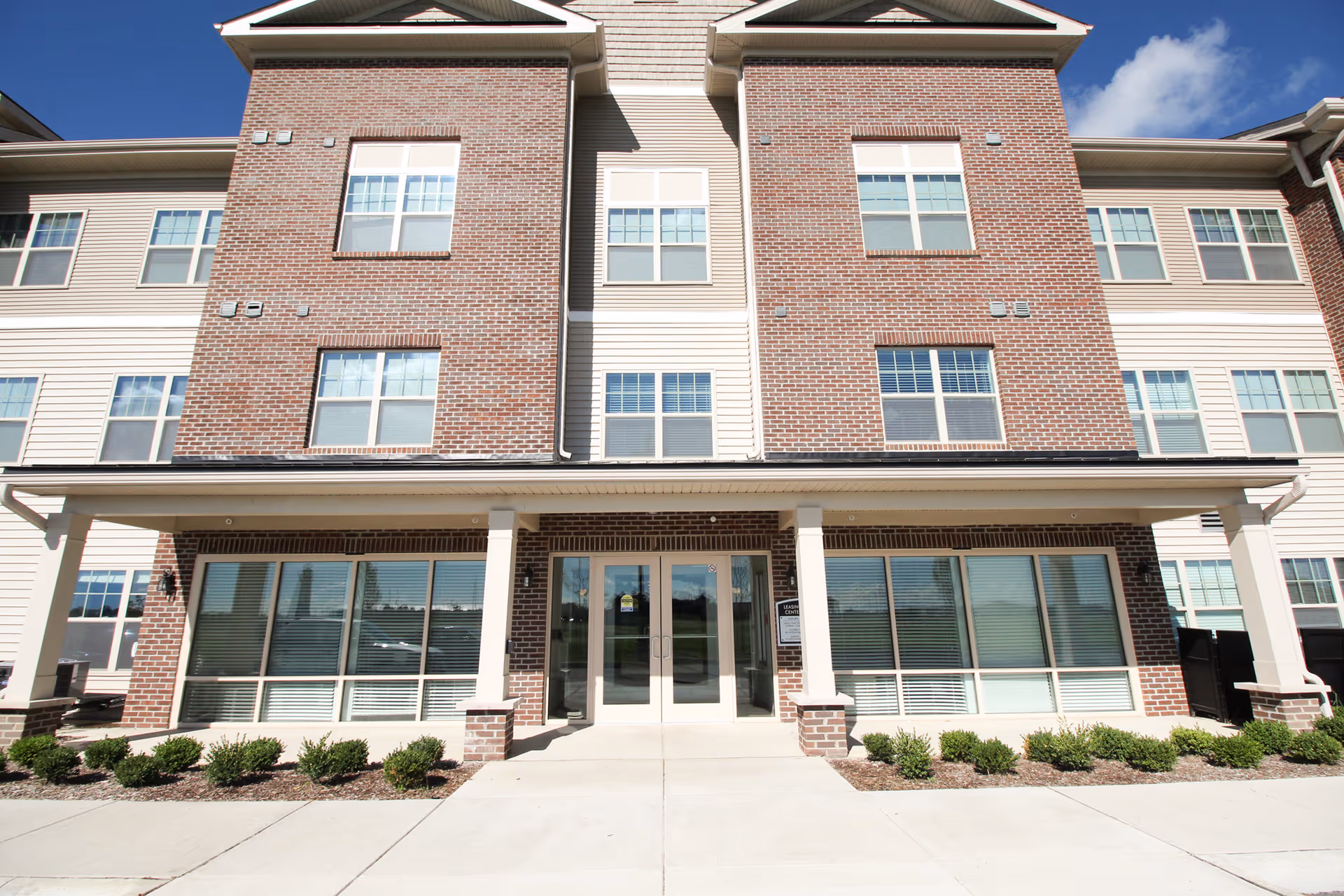 Front exterior view of a three-story senior living facility building with brick and siding facade, large windows, and a covered entrance with double glass doors. Small shrubs line the walkway leading to the entrance under a clear blue sky.