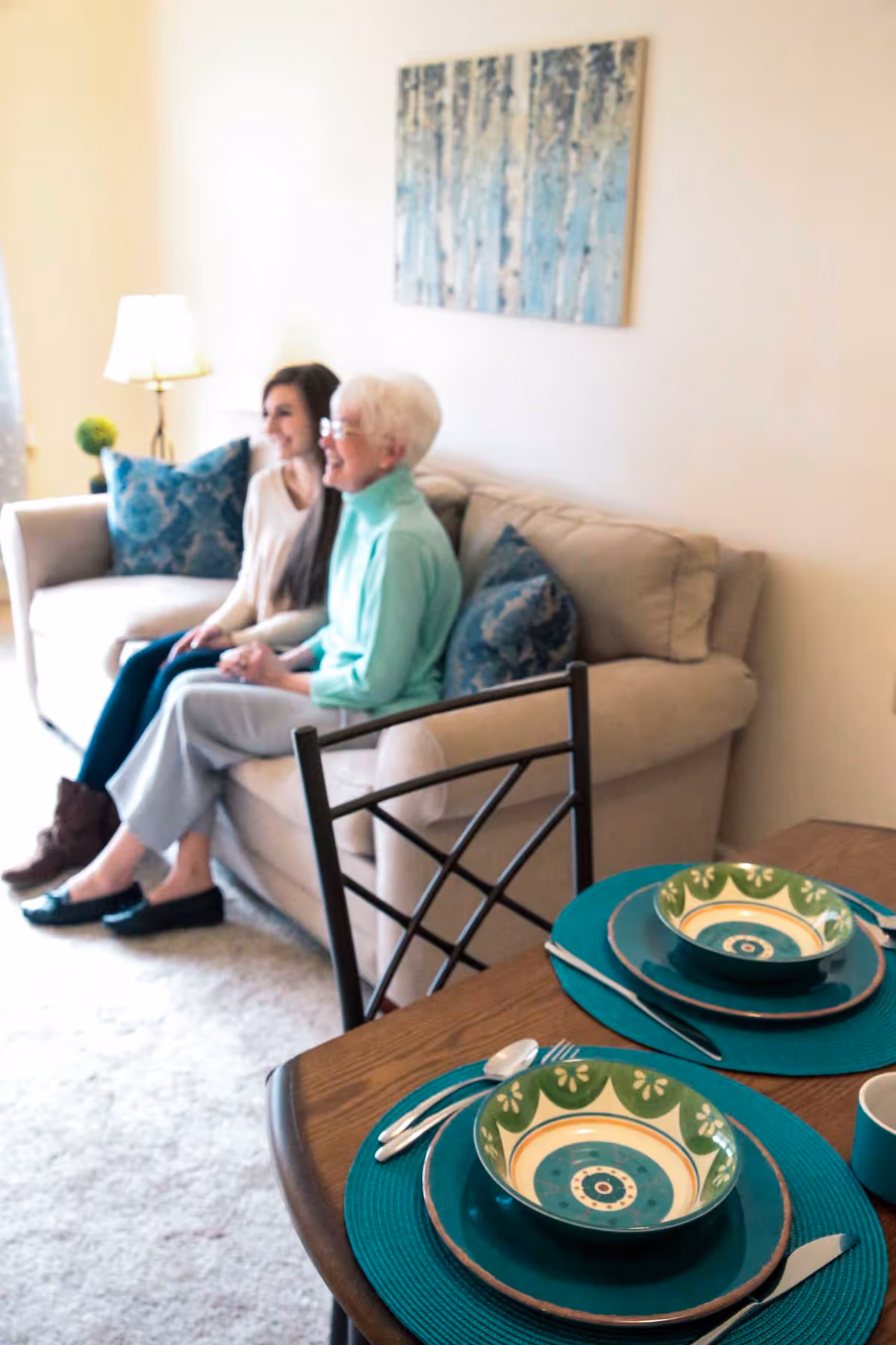 Two women sitting on a beige couch in a living room with blue patterned pillows and a painting of birch trees on the wall. In the foreground, a dining table is set with green and blue patterned bowls, plates, and silverware on teal placemats.