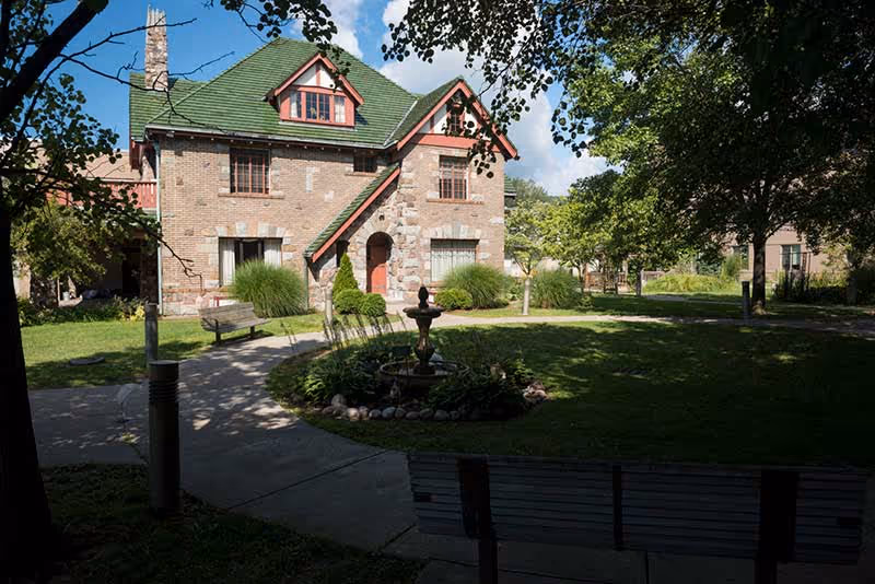 Brick two-story building with a green roof facing a shaded courtyard with a fountain, walkways, and benches.