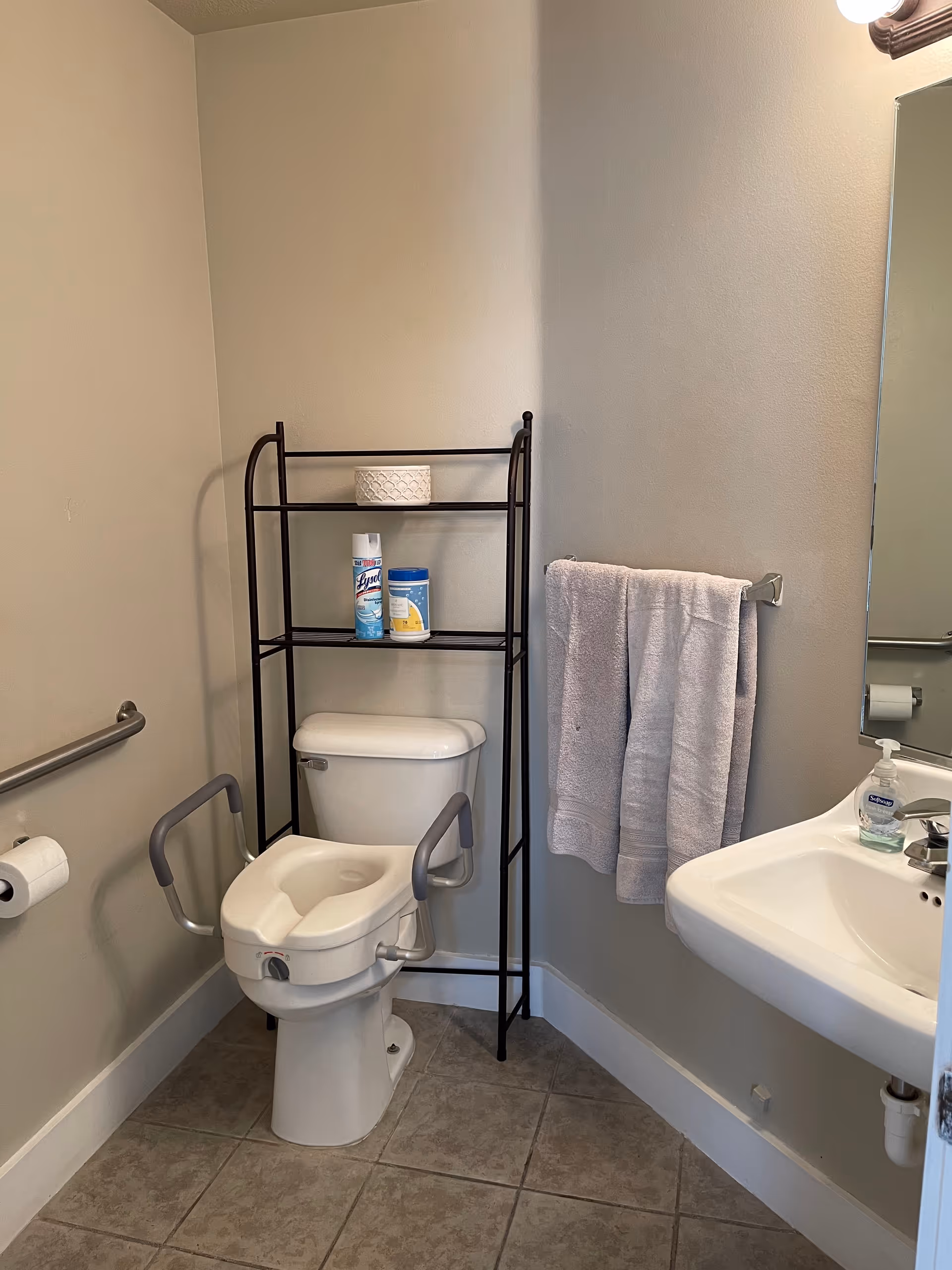Small accessible bathroom with a raised toilet with grab bars, a shelving unit holding cleaning supplies, a towel on a rack, and a wall-mounted sink.