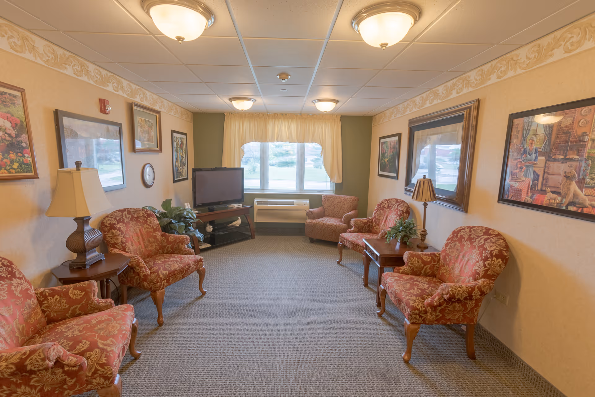 A cozy living room in a senior living facility with six upholstered armchairs arranged along the walls, two wooden side tables with lamps and plants, a flat-screen TV on a stand near a window with cream-colored curtains, and framed artwork on the walls.