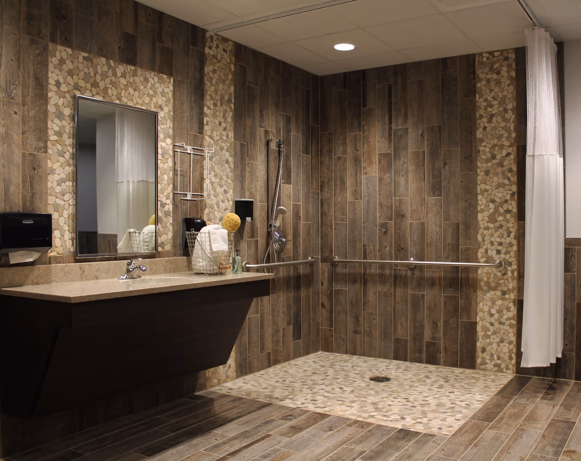 Accessible bathroom with a wall-mounted sink and mirror, grab bars and a walk-in shower with pebble tile floor and wood-look wall tiles.