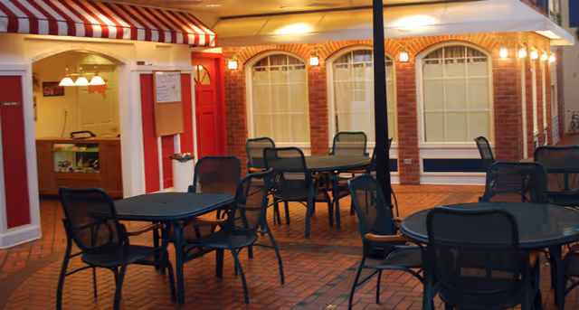 Outdoor patio area with multiple black metal tables and chairs on a brick floor, adjacent to a building with red brick walls and large windows covered with curtains. The building has a red door and a striped red and white awning above a counter window. Warm lights illuminate the area.