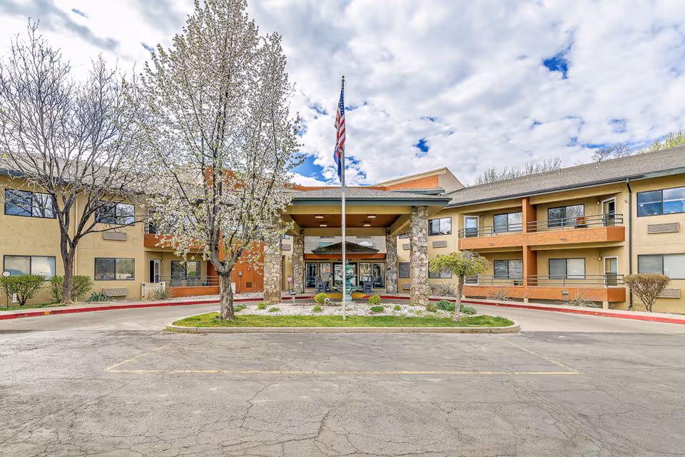 Front exterior view of Solstice Senior Living at Mesa View building with a covered entrance supported by stone pillars, an American flag on a flagpole in front, and trees with some greenery around the entrance. The sky is partly cloudy.