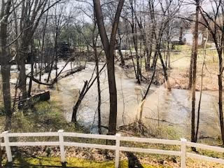 A view of a wooded area with leafless trees and a flowing creek or river. In the foreground, there is a white wooden fence and some grass. In the background, houses and other structures are partially visible through the trees.