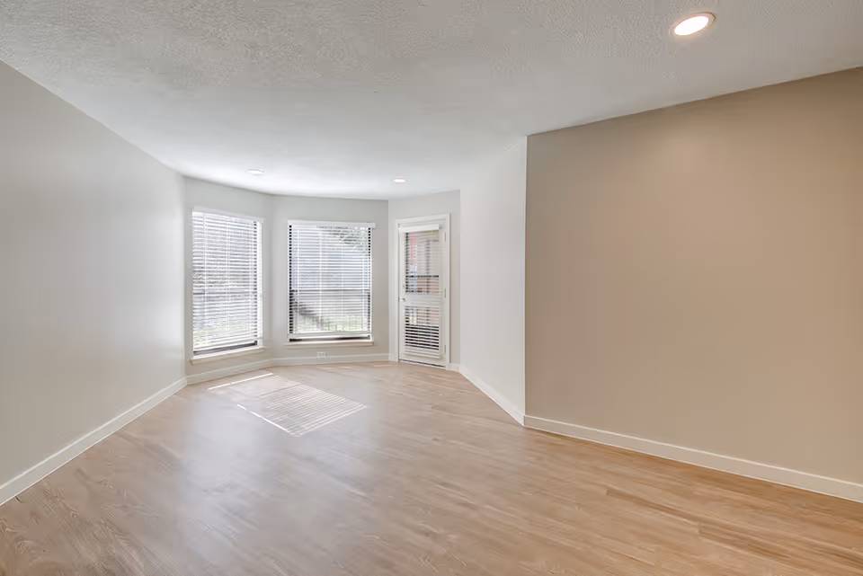 Empty room with light wood flooring, beige walls, and three large windows with white blinds allowing natural light to enter. There is a glass door with blinds on the right side of the windows. The ceiling has recessed lighting.